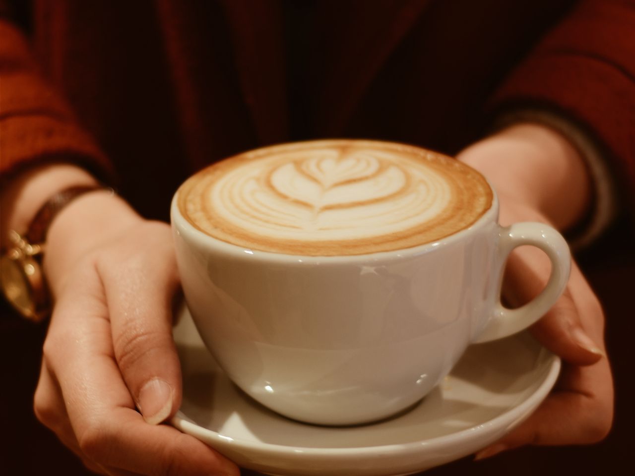 Two hands cradling a latte with feathered rosetta art in a pale cup